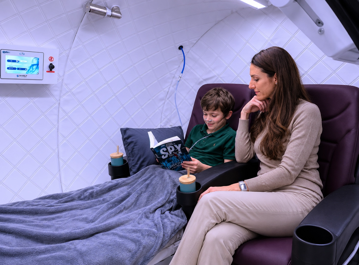 Mutter mit Sohn in einer HBOT Kammer mit Maske und Trinkbecher. Die Kammer bietet für zwei Menschen Platz.